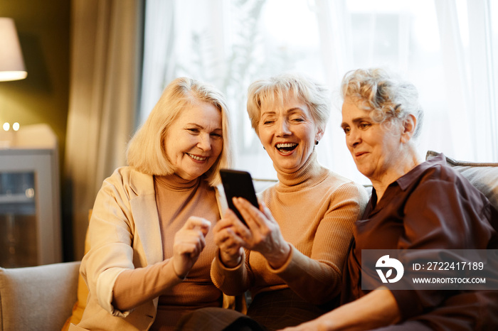 Group of three happy aged Caucasian women sitting together in living room against window and using smartphone