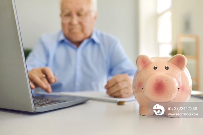 Piggy bank standing on desk with blurred senior man using laptop to pay bills online in background