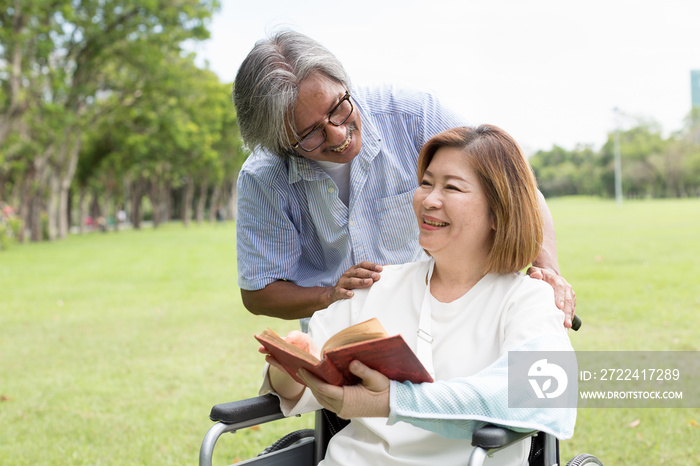 Asian elderly husband takes care his elderly sick wife while sitting on a wheelchair with injured arm wrapped in cast in the park. Asian senior couple spending time and relaxing together outdoor