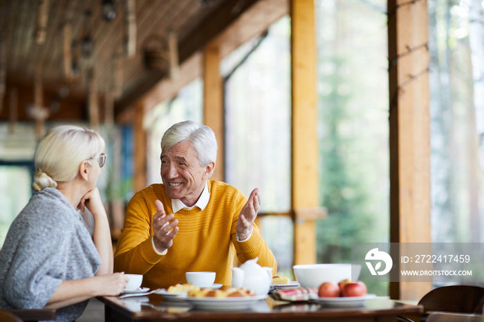 Senior grey-haired man in casualwear talking to his wife while both sitting by table in cafe