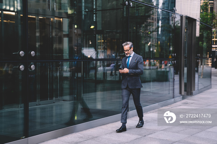 Elegant middle aged businessman tapping on mobile and walking down street