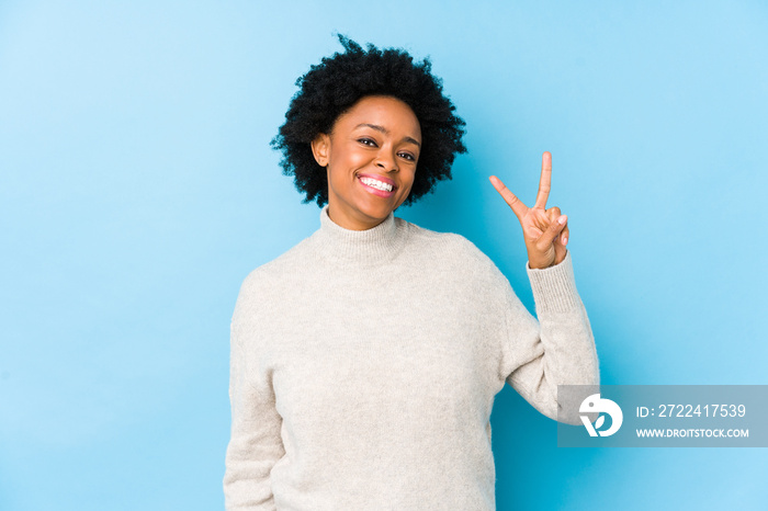 Middle aged african american woman against a blue background isolated showing victory sign and smiling broadly.