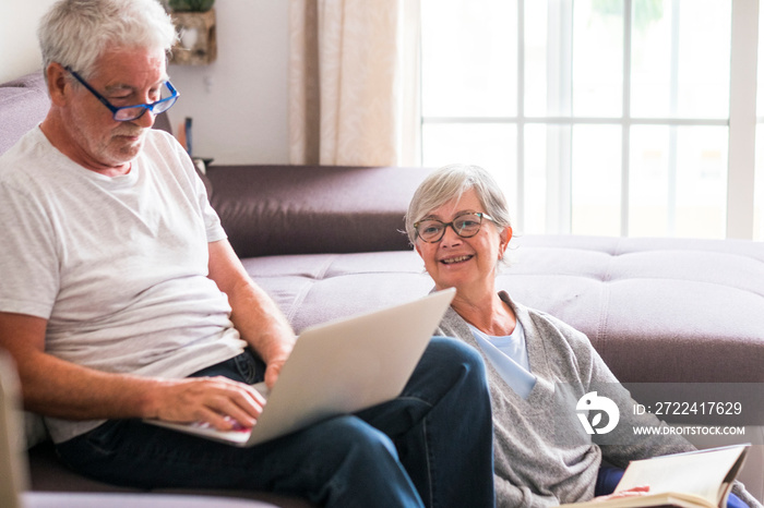 couple of seniors at home - man with laptop and glasses sitting at the sofa - woman sitting on the grund reading a book - indoor and love concept