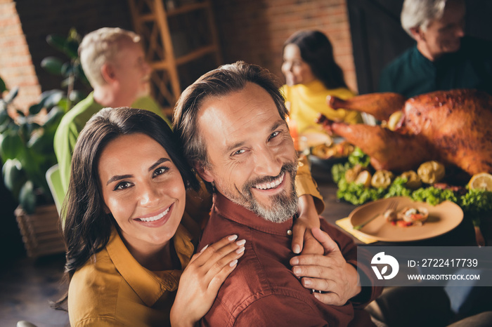 Closeup photo of full family gathering bearded husband wife cuddle beaming smiling hold hands finish eat meal sit served dinner big table turkey generation in evening living room indoors