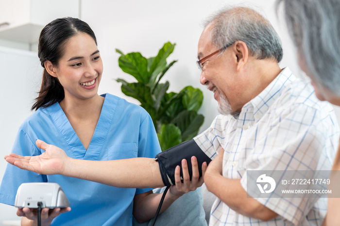 An elderly man having a blood pressure check by his personal caregiver with his wife sitting next to him in their home.