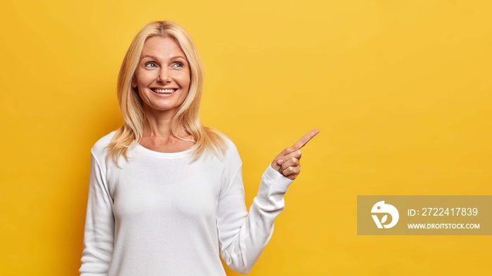 Happy senior woman shows copy space against yellow background smiles pleasantly and has dreamy expression demonstrates vacant advertising spot wears white jumper recommends good offer or discount