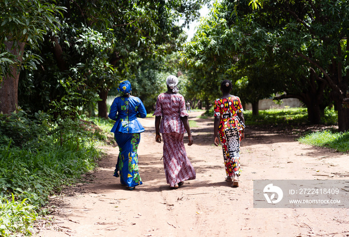 Rear view of three African women walking long distances on a dirt road due to a lack of transport links; missing infrastructure concept