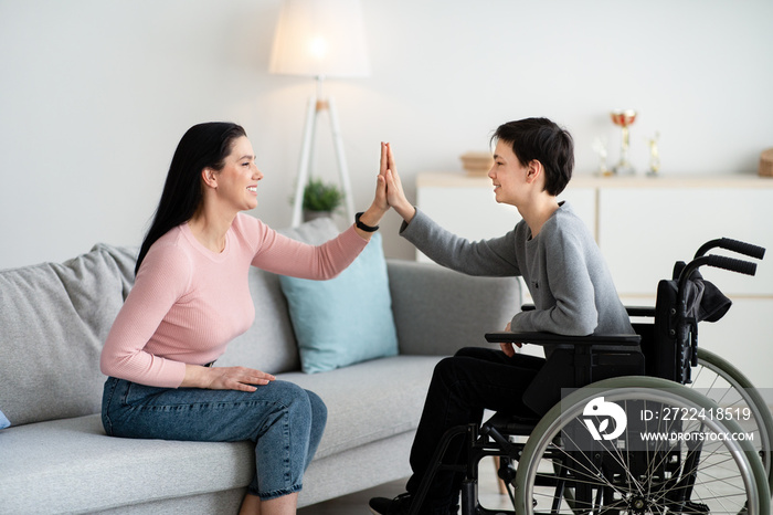 Happy teenage boy in wheelchair high fiving his mother at home, side view