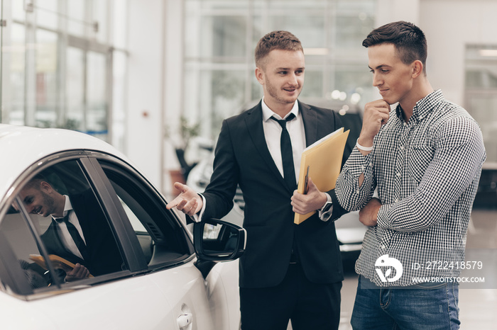 Professional car dealer showing cars on sale to his male customer, working at the auto dealership salon. Man looking thoughtfully at the car he is examining. Handsome man buying new automobile