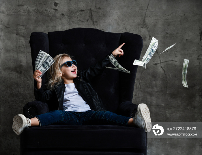 Happy smiling rich kid girl millionaire sitting in expensive armchair with a bundle of money dollars and euro cash bills