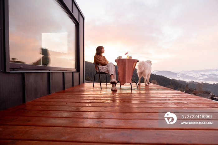 Woman with her dog resting on terrace of tiny house in the mountains, enjoying beautiful landscape during sunrise. Concept of small modern cabins for rest and escape to nature