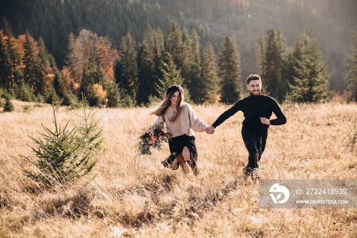 Stylish model couple in the autumn mountains. A young guy and a girl run along the slope against the background of the forest and mountain peaks at sunset. Girl holds in her hands a bouquet of flowers