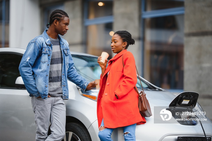 Happy young adult african american man and smiling woman charging electric car.