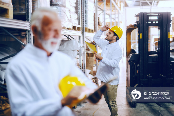 Blue collar employees working in warehouse. In foreground senior adult worker using tablet. Selective focus on younger worker checking on goods in warehouse.