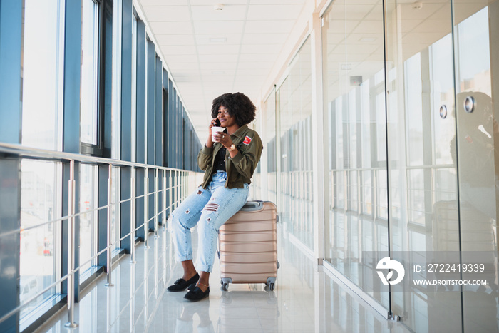 Portrait of happy young african american woman sitting on suitcase and talking with mobile phone at the station