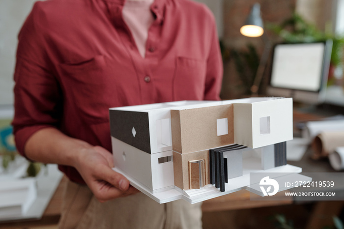 Hand of young female architect or engineering designer in casualwear holding paper model of new house while standing in office