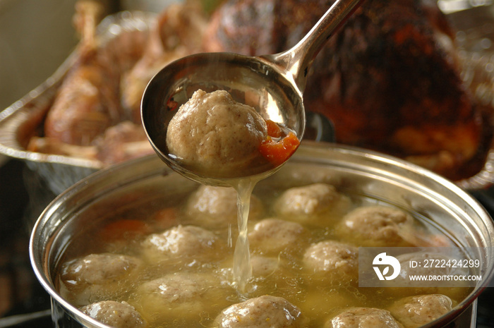 Large pot of chicken soup with fluffy matzah balls floating on the surface and roast turkey in the background.