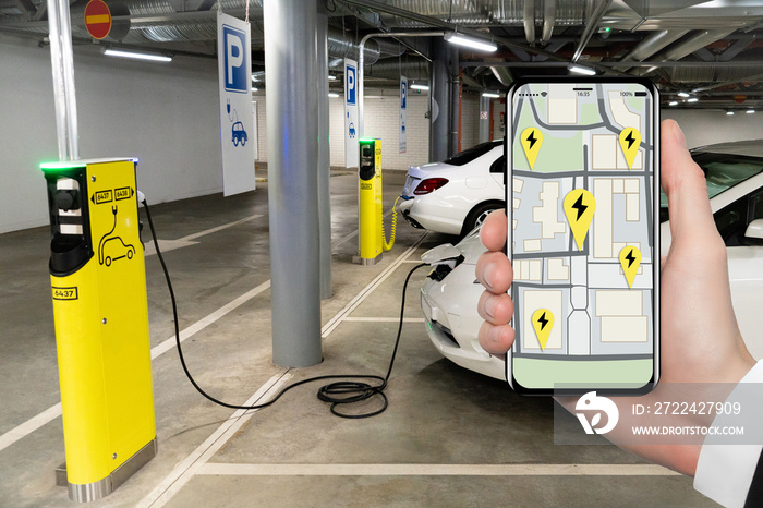 A hand with a phone on the background of a charging station for electric cars. On the screen is a map with the location of the charging stations.