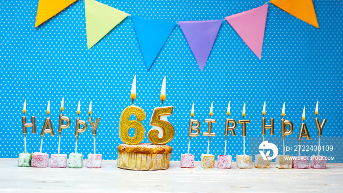 Happy birthday from the letters of candles number 65 on a blue background with polka dots white copy space. Happy birthday muffin with burning golden color candle for sixty five years anniversary