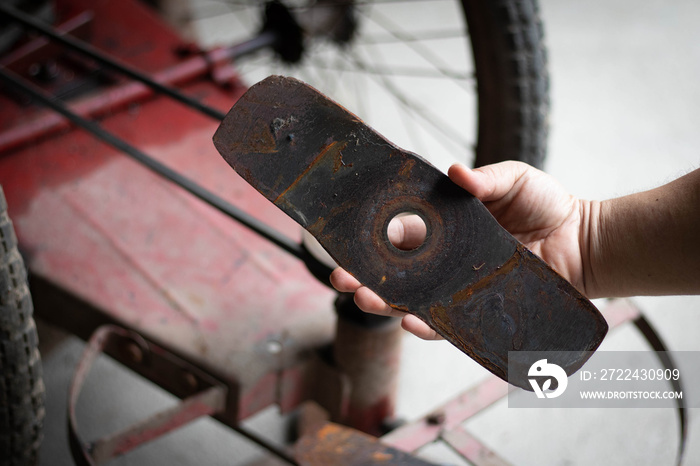 Man’s hand holding a blade with 2 lawn mowers, rusted blades. In the background is a lawn mower.