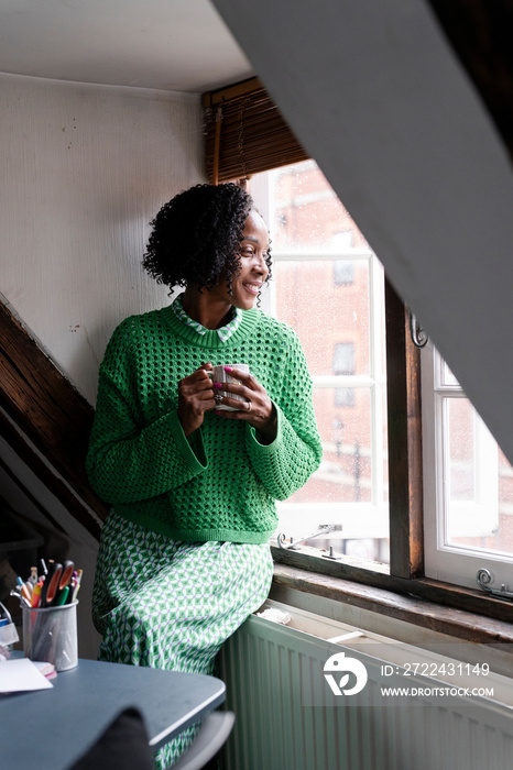 Smiling businesswoman holding cup of coffee while looking through window