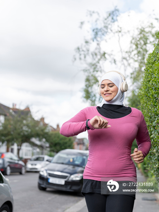 UK,Sutton,Woman in headscarf and headphones jogging and looking at smart watch