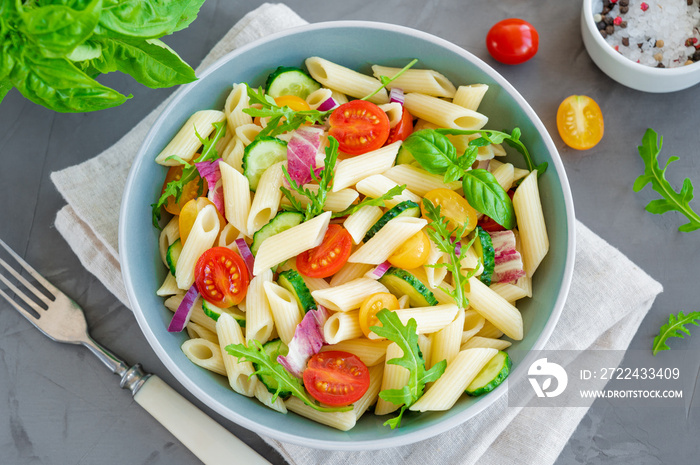 Healthy vegan pasta salad with tomatoes, cucumbers, red onions and arugula in a bowl on a dark gray background. Copy space.