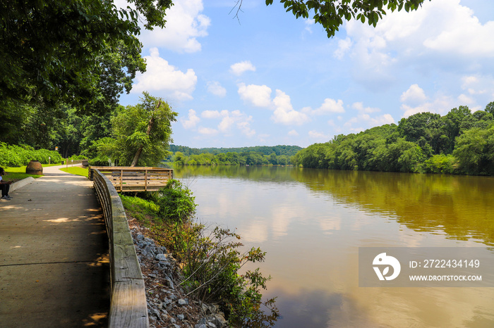 a shot of the still waters of the Chattahoochee river with lush green trees reflecting off the water with a boardwalk with wooden fence over the water, blue sky and clouds at Roswell Riverwalk