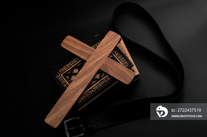 Spare the rod, spoil the child, lost innocence and children abuse concept with religious wood cross, stack of books and leather belt isolated on dark black background