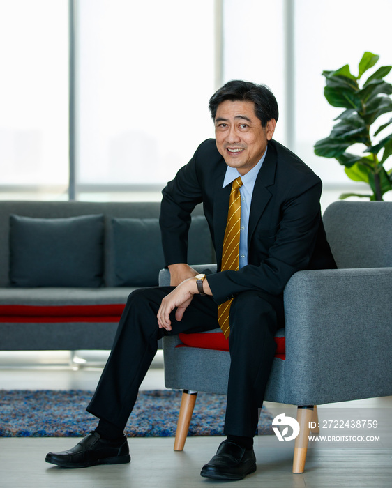 50s Asian senior executive businessman entrepreneur wearing formal suit with luxury necktie, smart sitting on sofa in indoor office, smiling with confidence, success and gladness with copy space.