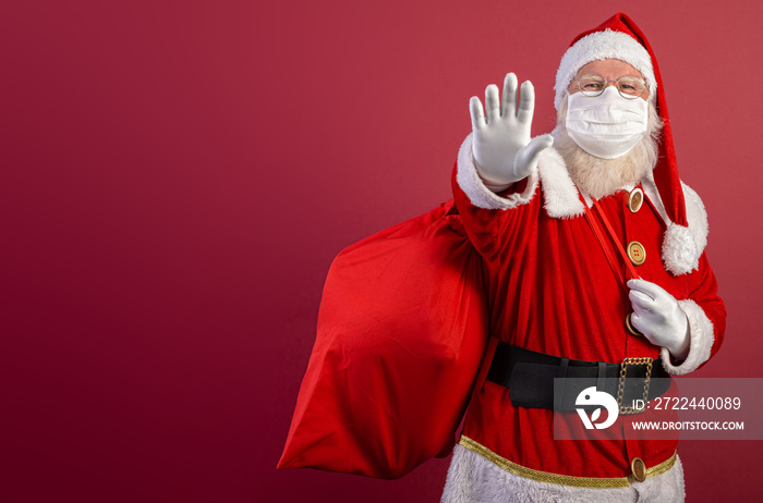 Real Santa Claus on red background, wearing protective mask and with an open hand in front making a pandemic stop sign. Christmas with social distance. Covid-19