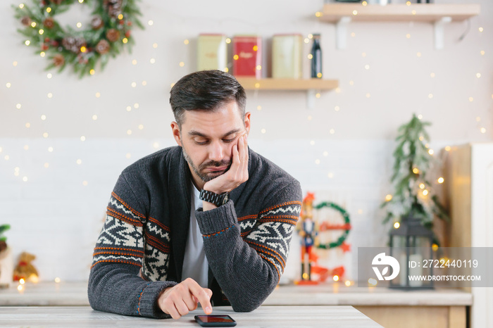 A man with a beard is bored and is alone at home for Christmas, looking at the phone in depression