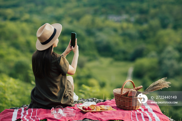 Young girl with hat making photo on smartphone at beautiful landscape while resting at summer picnic