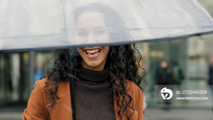 Portrait of beautiful cute flirting brunette woman with curly hair attractive girl tourist with transparent umbrella posing on street in city in rainy weather enjoying rain looking at camera smiling