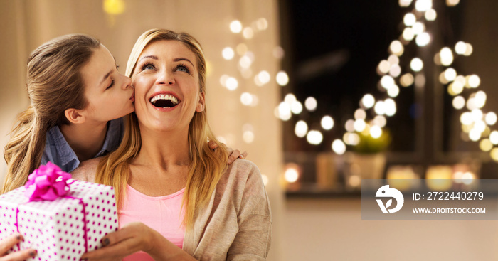 people, holidays and family concept - happy daughter giving birthday present and kissing her mother at home over garland lights background