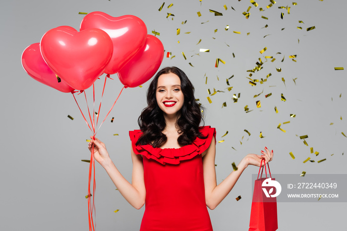 Happy woman holding red heart-shaped balloons and shopping bag near falling confetti on grey