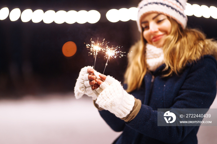 Sparklers in hands. Beautiful young woman in the winter time holding a sparkler in hand in the forest. Happy cute girl in knitted hat posing with sparkler. Festive garland lights. Christmas, new year.