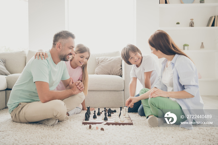 Photo portrait of little brother and sister sitting with parents on carpet playing chess together smiling happy