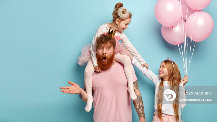 Young ginger bearded father left alone with two children, has shocked expression, can not calm down naughty daughters who tickle and laugh, being in good mood on party for kids. Studio shot.