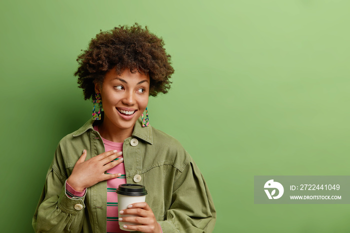 Happy sincere Afro American lady concentrated positively aside smiles broadly dressed in stylish clothing drinks takeaway coffee isolated over vibrant green background with copy space aside.