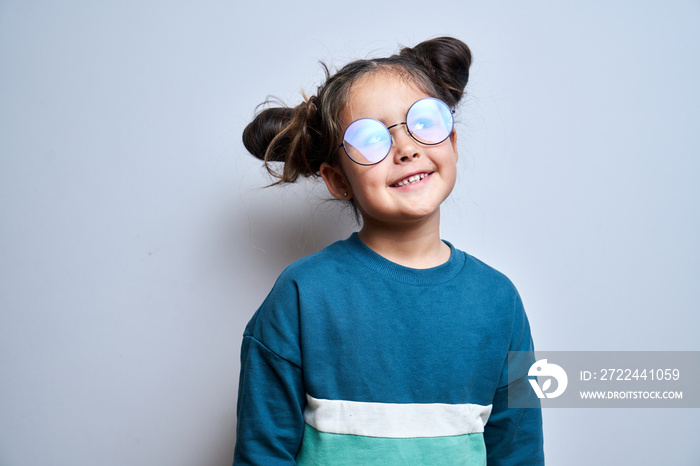 Portrait of smart dreamy little girl in glasses and funny hairstyle smiles and looks away isolated on white studio background, happy beautiful child