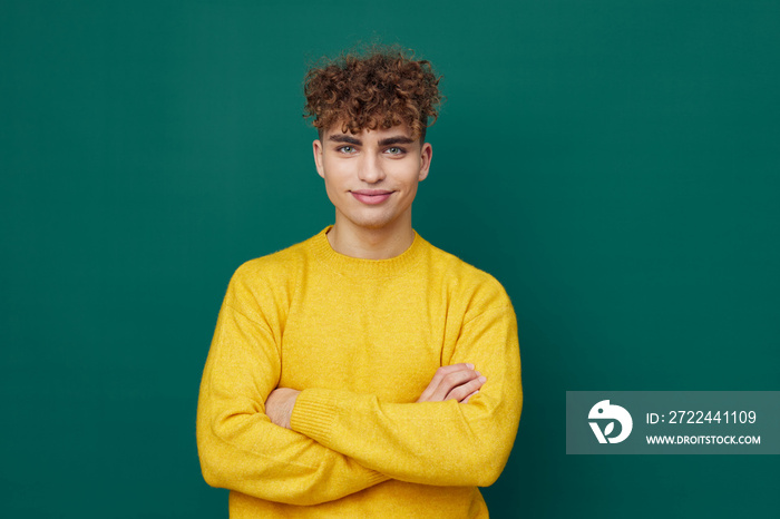 a handsome man stands on a green background in a yellow sweater and smiling pleasantly poses with his arms folded on his chest . Horizontal photo with empty space