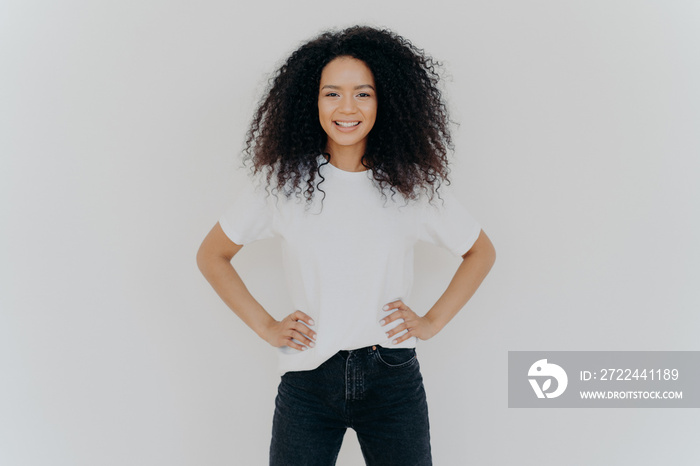 Photo of delighted curly woman keeps both hands on waist, smiles gently, has slim figure, wears white t shirt and black jeans, being in good mood, stands self assured against white background