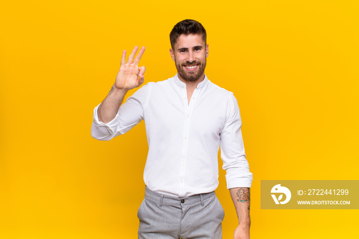 young handosme man feeling happy, relaxed and satisfied, showing approval with okay gesture, smiling against flat color wall