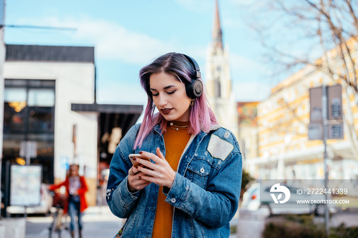 Beautiful girl with purple hair and headphones on the city street. Looking at phone.