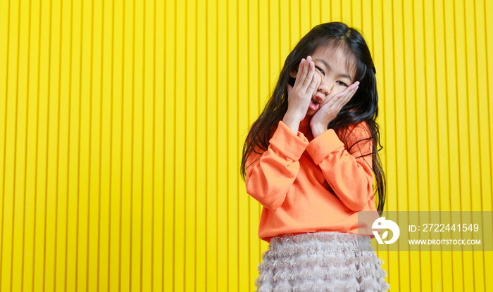 Portrait studio shot Asian playful cheerful funny face young primary school girl daughter in casual outfit holding hands squeezing cheeks and mouth looking at camera on yellow stripe wall background