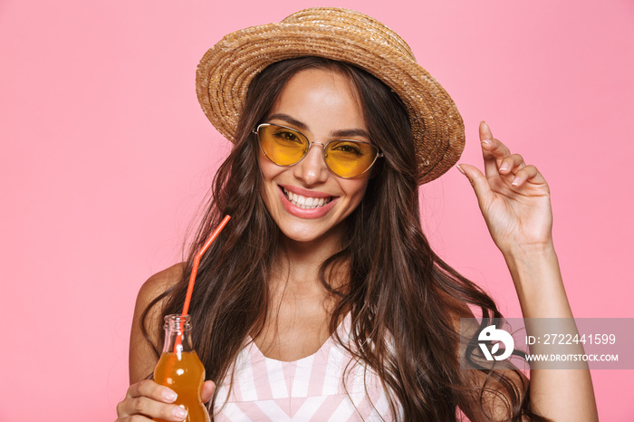 Photo closeup of fashion woman 20s wearing sunglasses and straw hat drinking soda from glass bottle, isolated over pink background