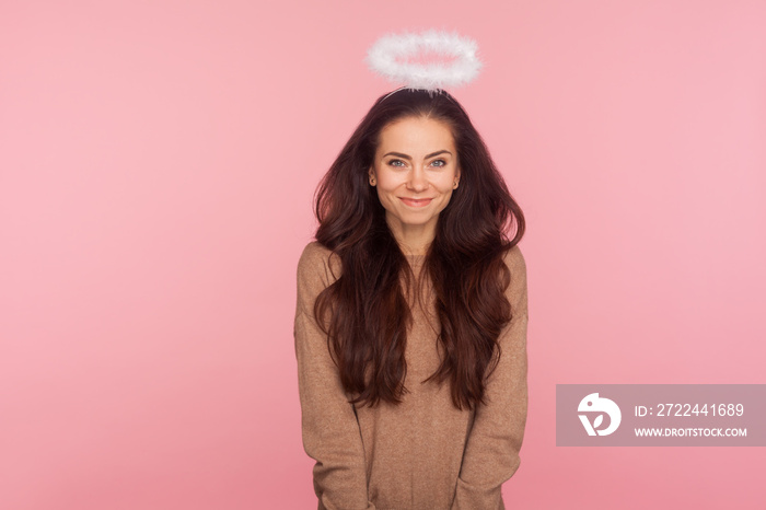 Portrait of lovely shy young woman with halo above head looking at camera with kind timid smile, expressing angelic beauty, sensuality and obedience. indoor studio shot isolated on pink background
