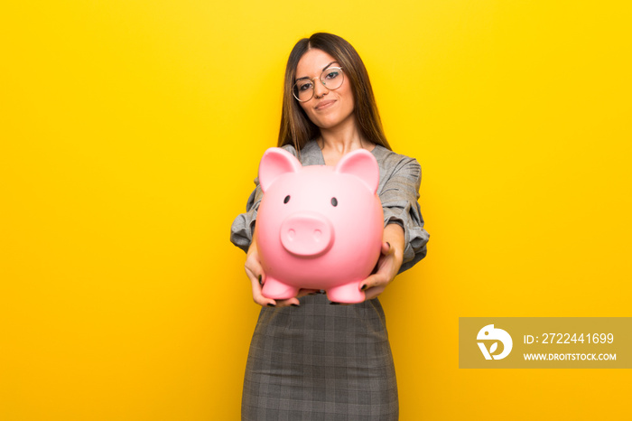 Young woman with glasses over yellow wall holding a piggybank