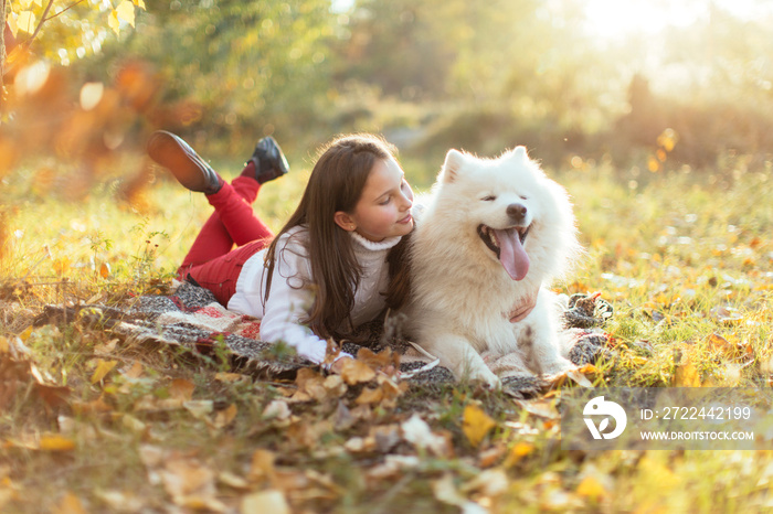 A little brunette girl with her white fluffy dog lies on autumn leaves in the park. Friendship and harmony in the family. Happy childhood.